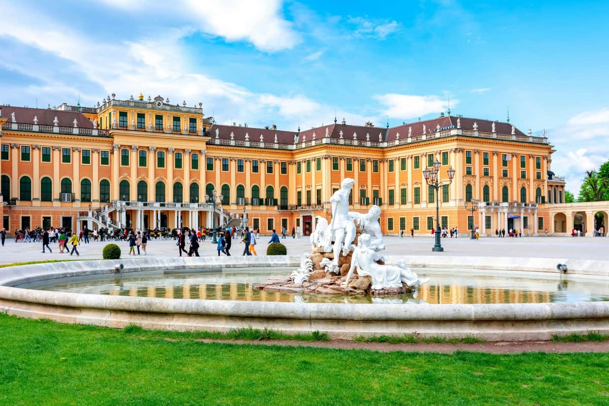A large historic building with a yellow facade, a decorative fountain in the foreground, and people walking around.