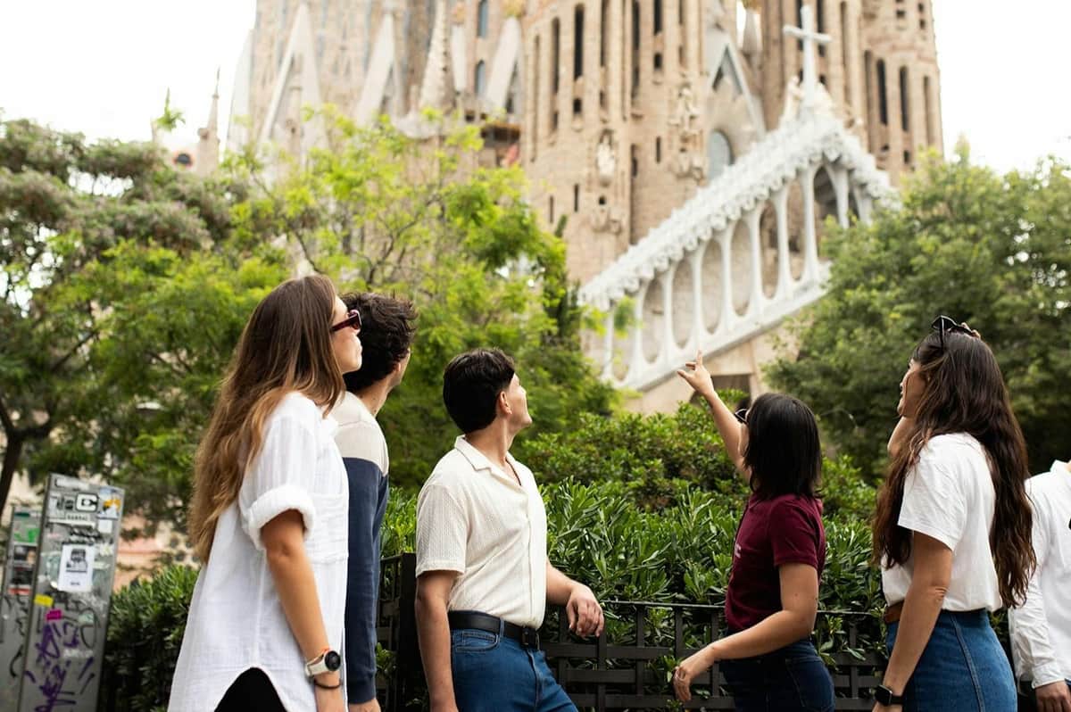 Barcelone : visite guidée + billets coupe-file pour le Parc Güell et la Sagrada Familia (Barcelone) Trois personnes se tiennent dans un parc, le regard tourné vers la Sagrada Familia en arrière-plan, au milieu de la verdure et d'éléments urbains.