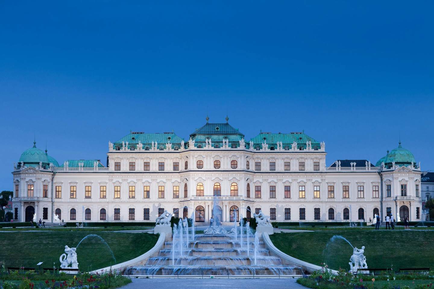 An ornate palace with green domes, fountains, statues, and gardens, set against a clear blue sky at dusk.