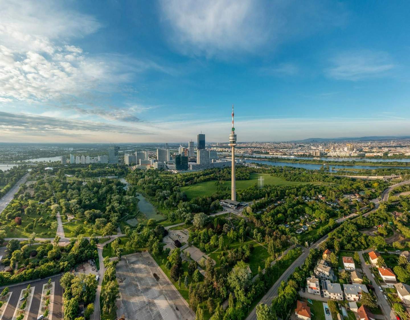 Danube Tower City Card (Vienna) Aerial view of a city with a tall tower, green parks, a river, and modern buildings under a blue sky with clouds.