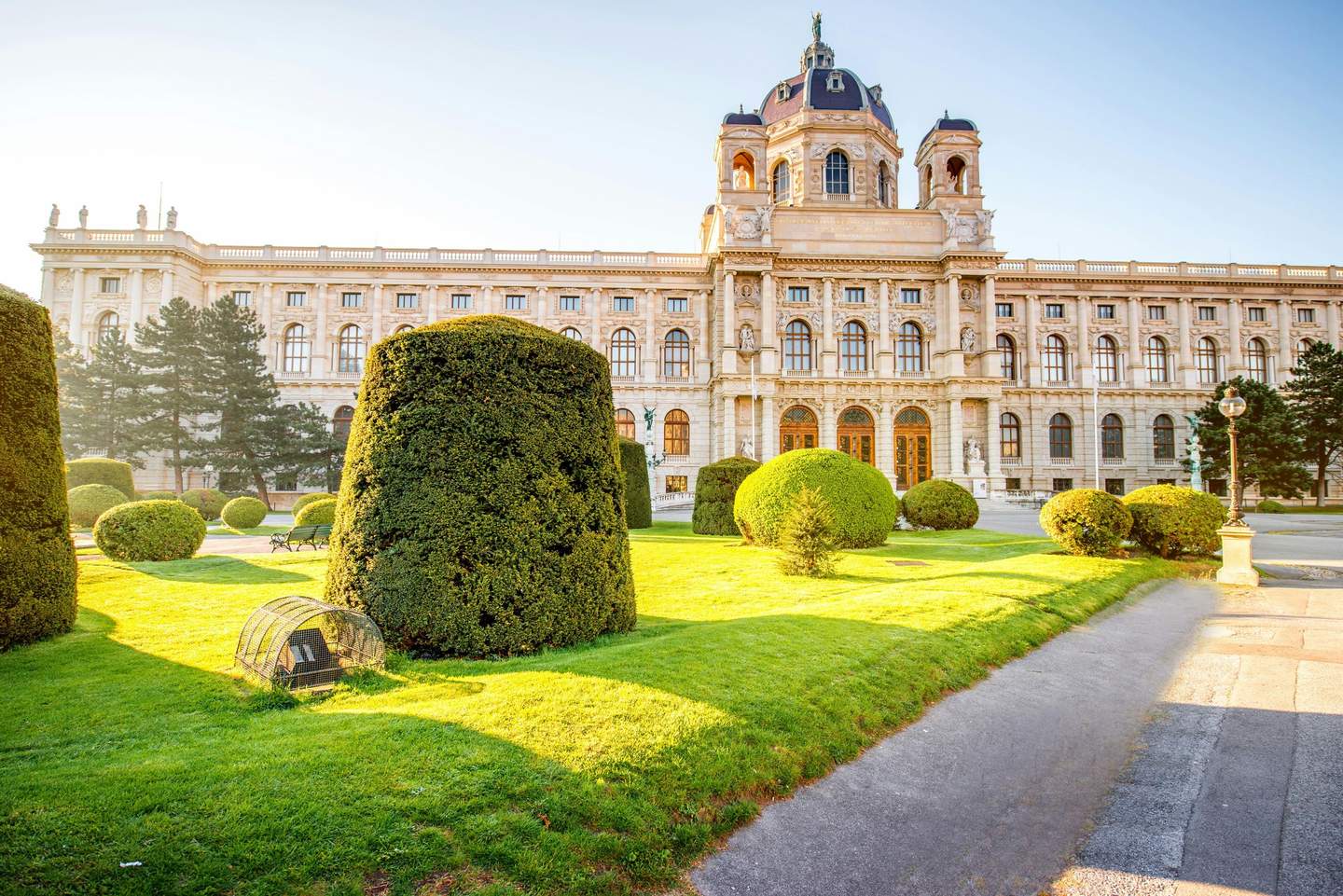 A large, historic building with arched windows, a dome, and neatly trimmed hedges in a sunlit garden area.