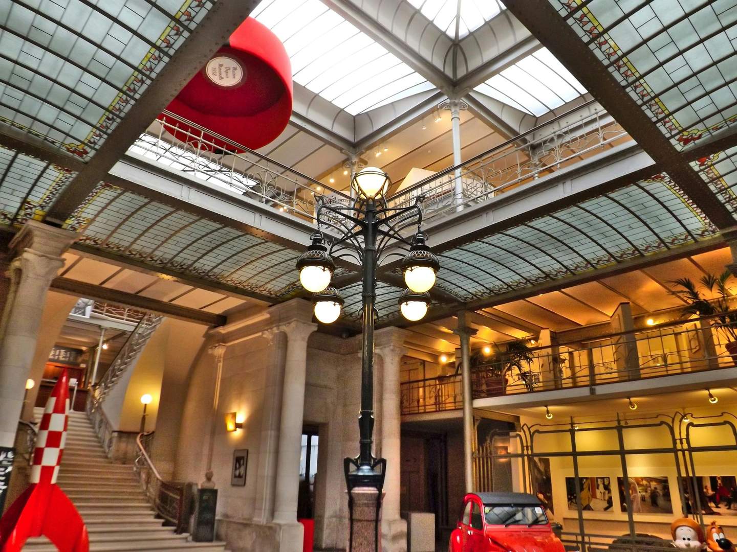 Interior of a vintage building with a glass ceiling, ornate railing, central lamp post, stairs, and a red car display near a gallery.