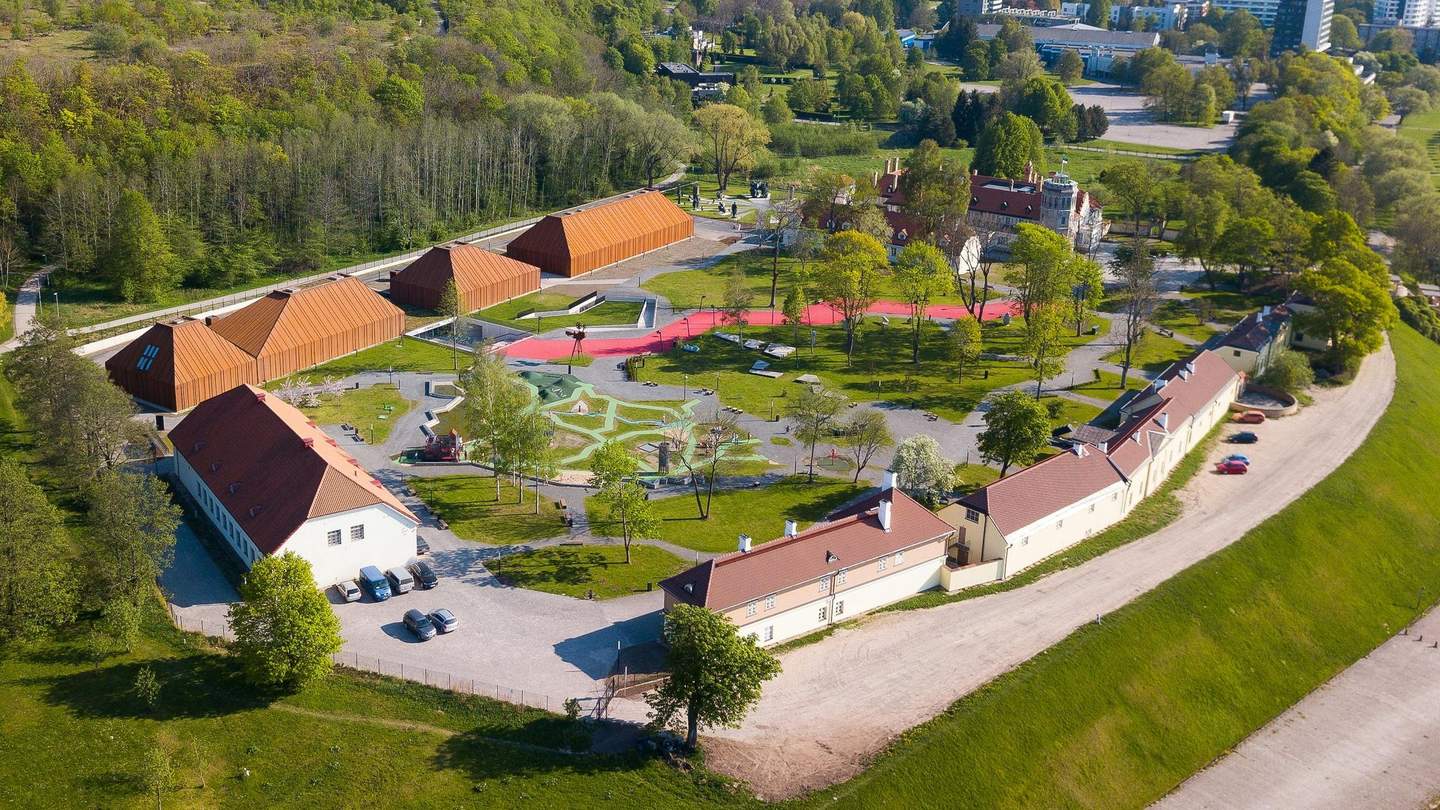 Aerial view of a park with pathways, playgrounds, brown-roofed buildings, parking lot, and surrounding greenery on a sunny day.