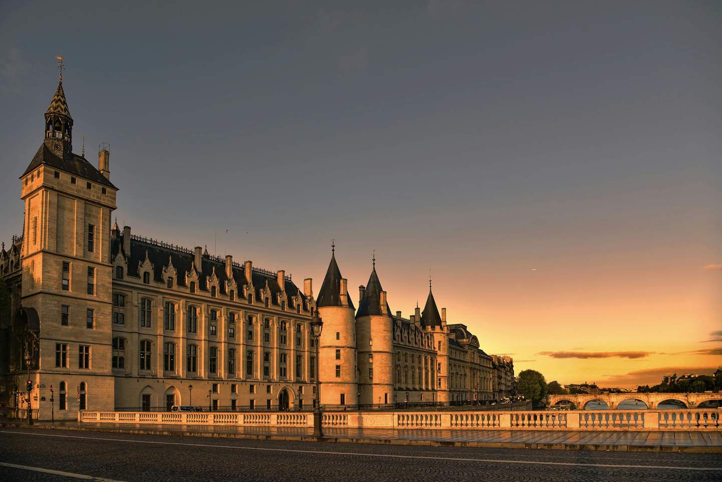 Conciergerie City Card (Paris) Historic building with tower and turrets along a river at sunset, with a bridge in the background and clear sky.