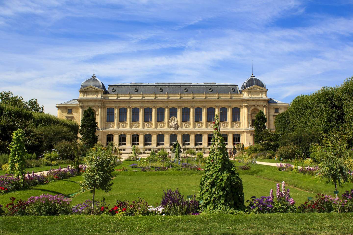 Ornate building with large windows and domed rooftops, framed by lush gardens and colorful flower beds under a blue sky.