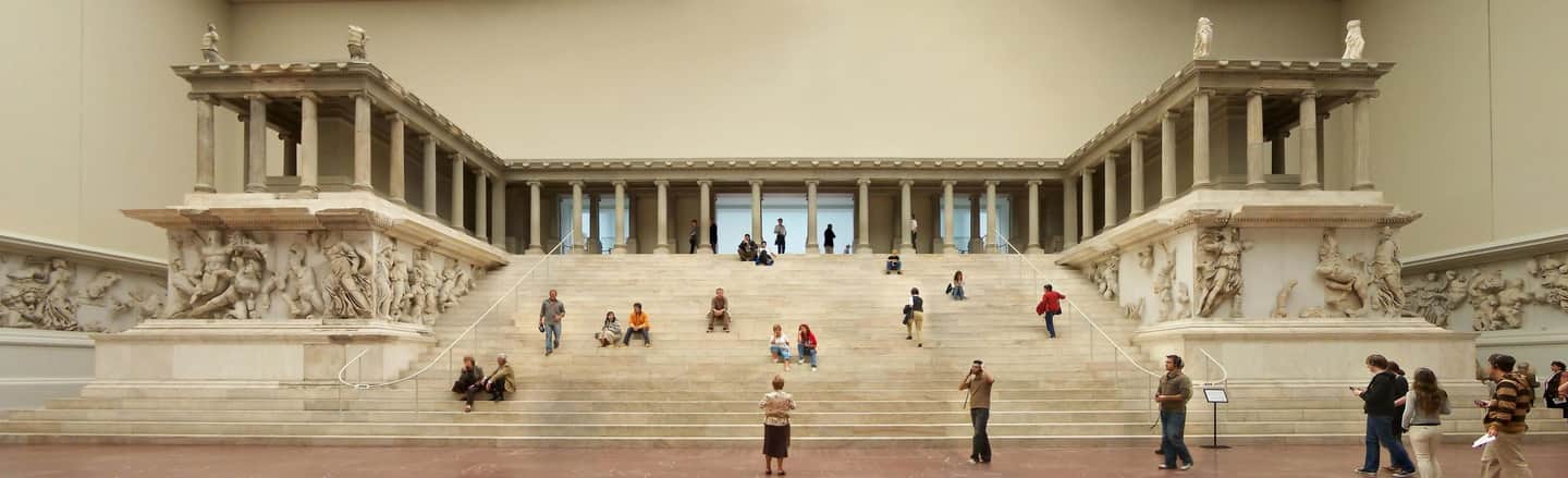 Pergamon Museum City Card (Berlin) A grand stairway flanked by large architectural sculptures and columns, with people walking, sitting, and observing the artwork.