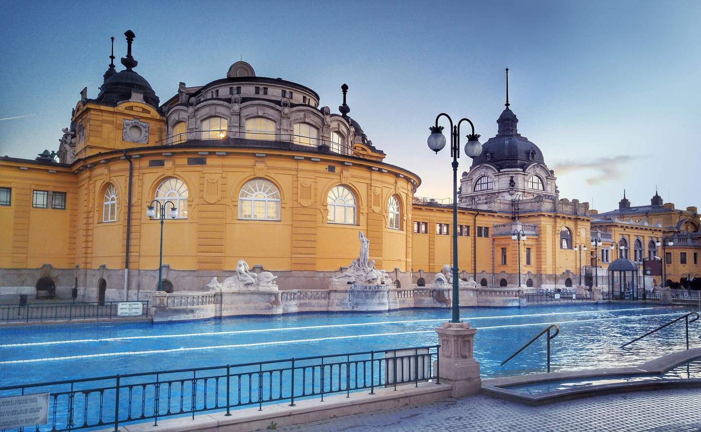Ornate yellow building with arched windows, dome-shaped roof, statues, and a pool in the foreground under a clear sky.