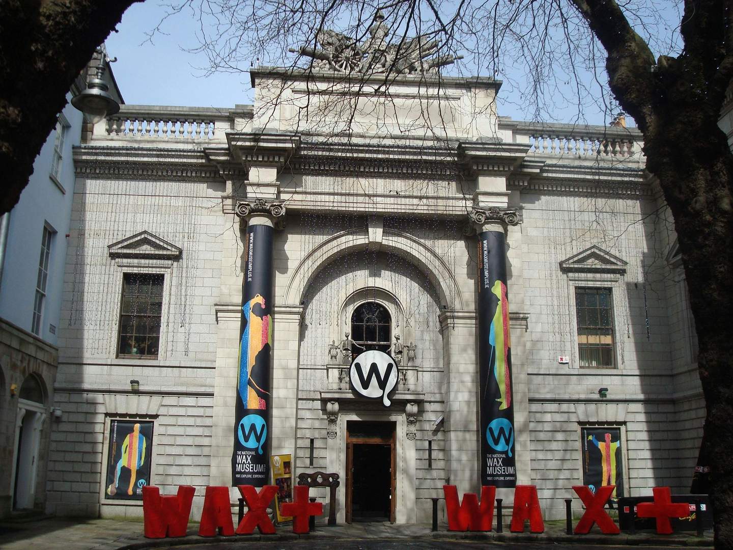 Front facade of the Wax Museum building with a stone exterior, large pillars, banners, and a "WAX+" sign.