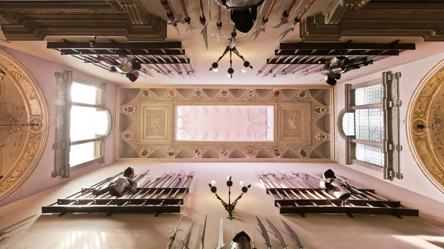 Ornate hallway ceiling with medieval armor suits and weaponry displayed on the walls, flanked by tall, arched windows.