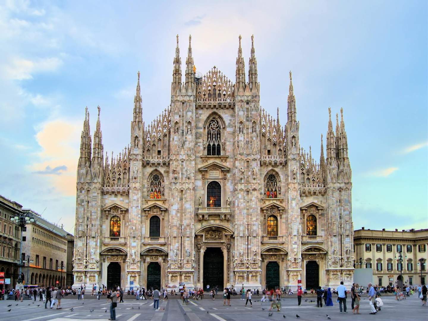 Duomo di Milano City Card (Milan) People gather in front of an ornate Gothic cathedral with intricate spires and stained glass windows under a blue and cloudy sky.