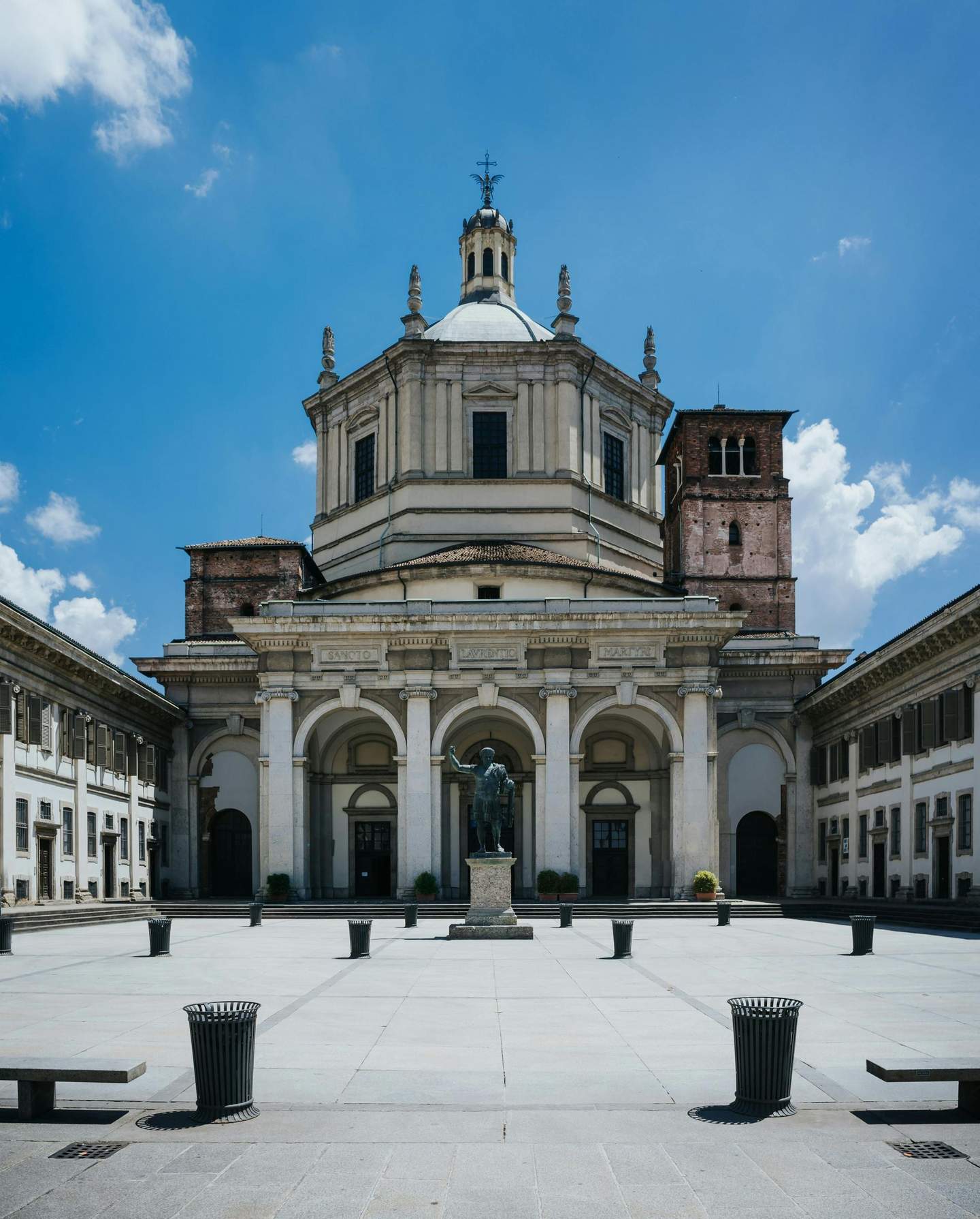 A historic building with a dome, arches, and a statue in the front courtyard, surrounded by columns and a clear blue sky.