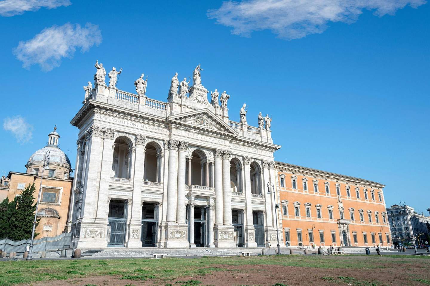 Historic church facade with statues, arched windows, and columns under a clear blue sky, adjacent to a dome and orange building.