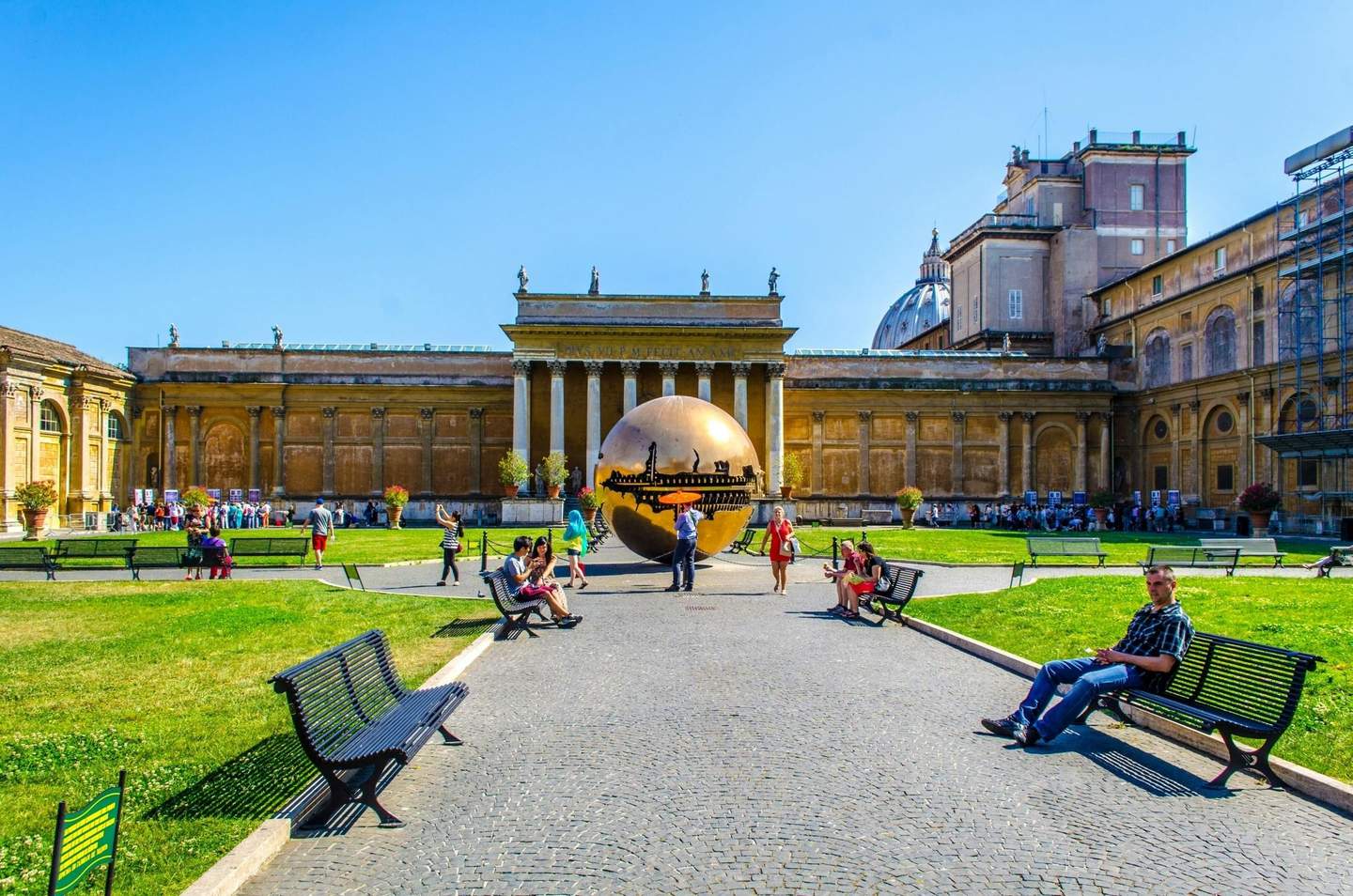 Tourists relax on benches in a courtyard with a large bronze sculpture and historic building under a clear blue sky.