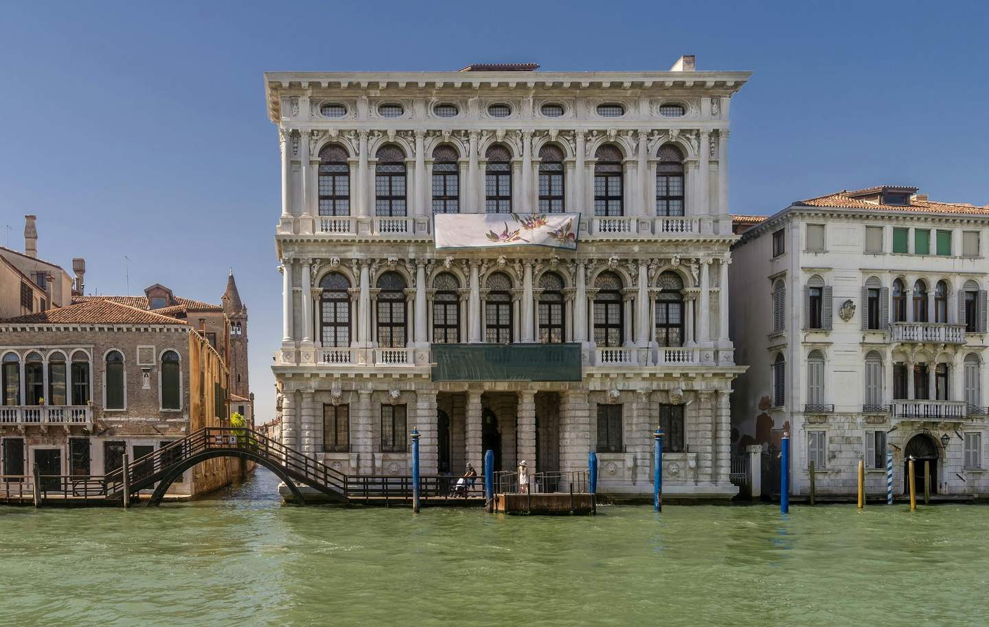 Targetes de la ciutat per a Museu Ca' Rezzonico (Venècia) Un edifici gran i ornamentat al costat d'un canal amb una pancarta, envoltat d'edificis més petits i una passarel·la de fusta sobre l'aigua.