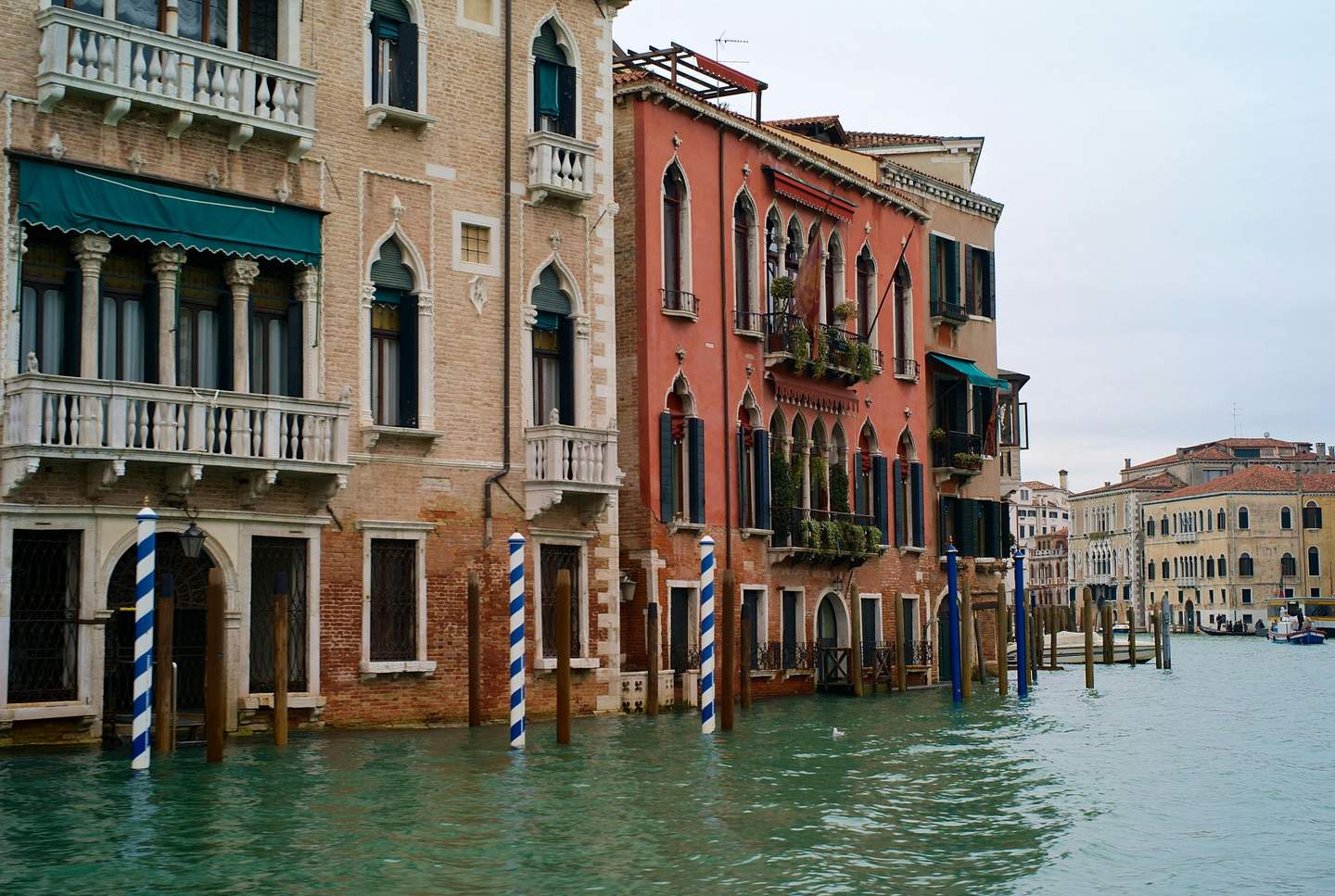 Mocenigo Palace-Museum City Card (Venice) Colorful buildings line a canal in Venice, with striped blue and white mooring poles, balconies, and arched windows.