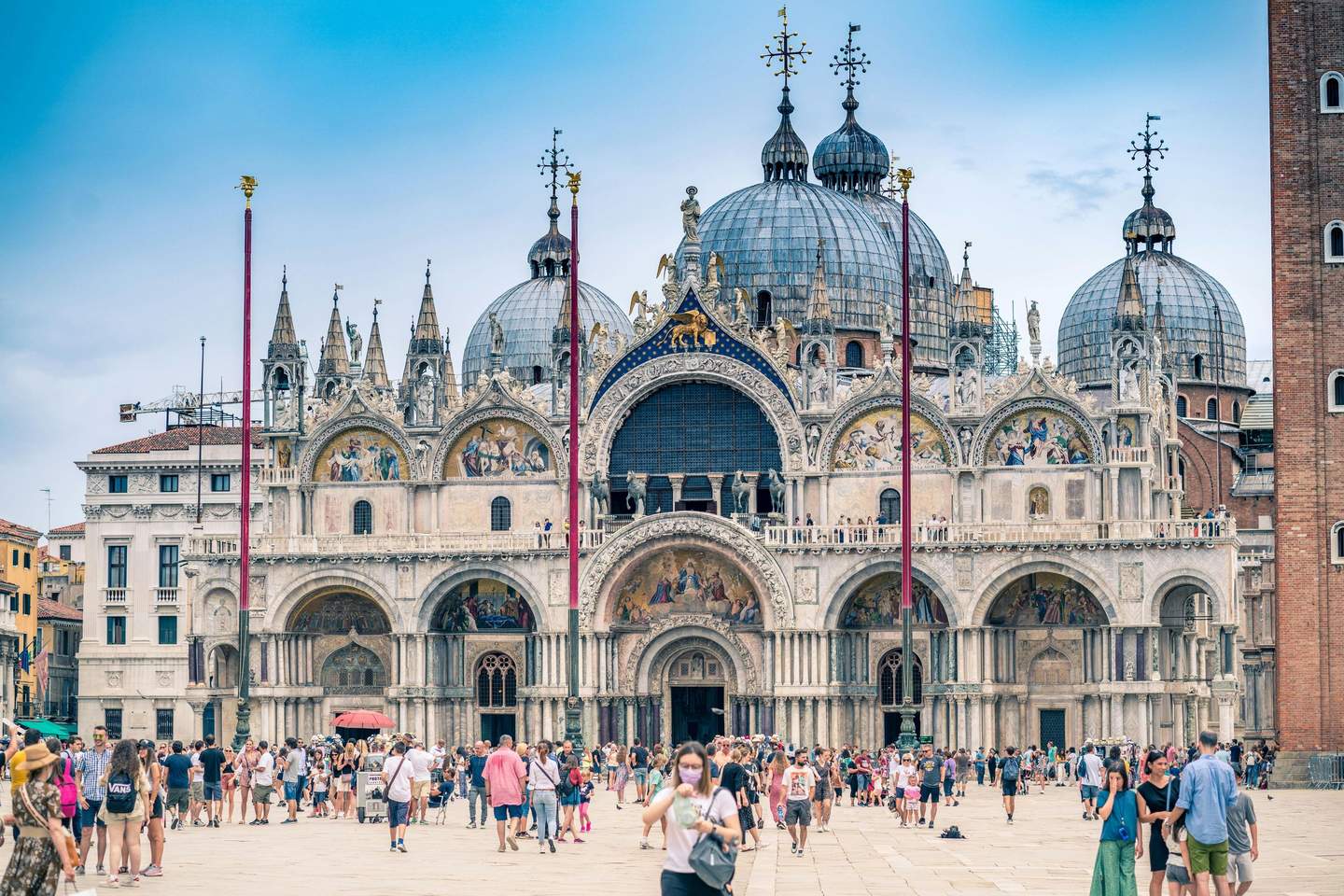Tourists walking and gathering in front of a large, ornate cathedral with domes and intricate facades under a clear sky.