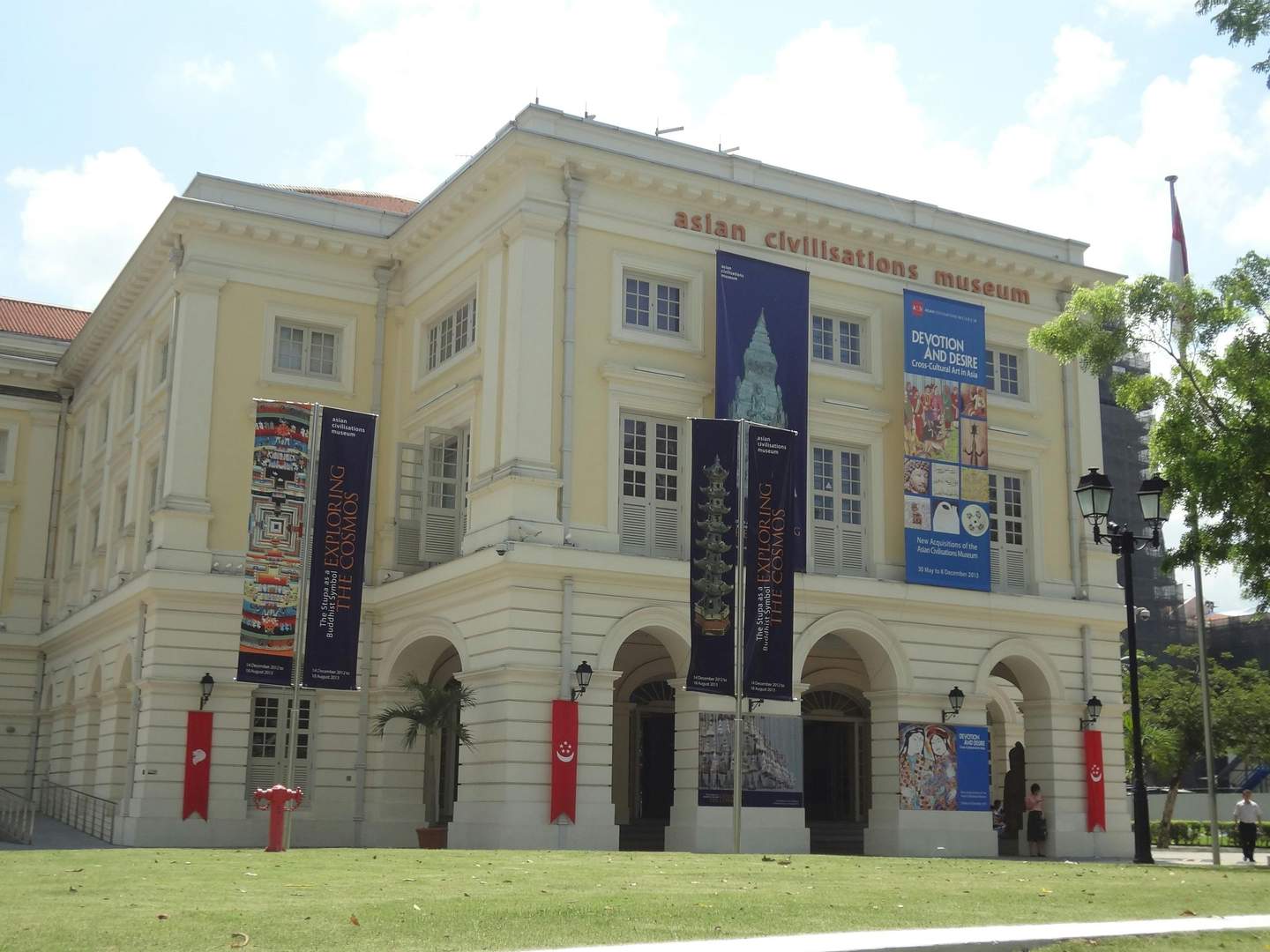 Un gran edificio amarillo con carteles de "Museo de las Civilizaciones Asiáticas" y pancartas para exposiciones, rodeado de césped y árboles.