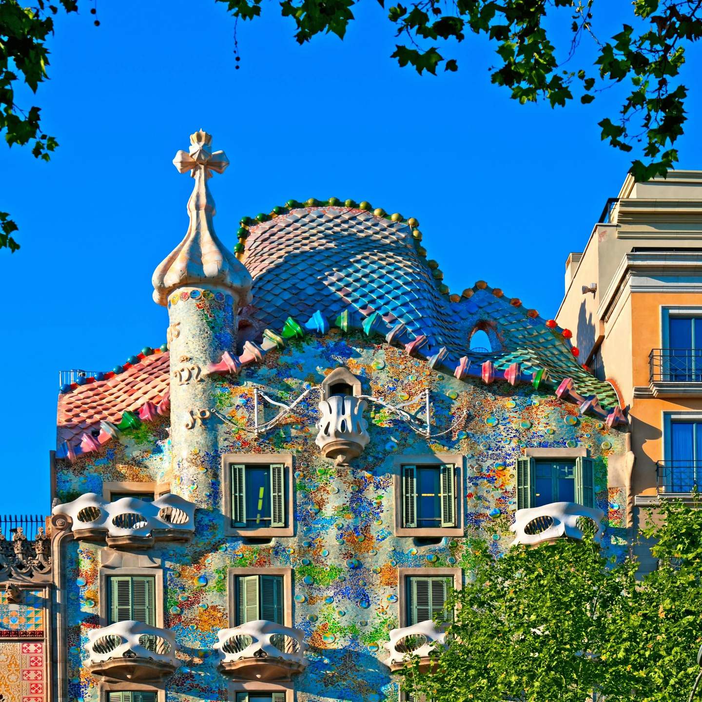 Un edifici de colors cobert de mosaics amb balcons corbats, persianes verdes i una torreta en forma de con, emmarcada per branques d'arbres.