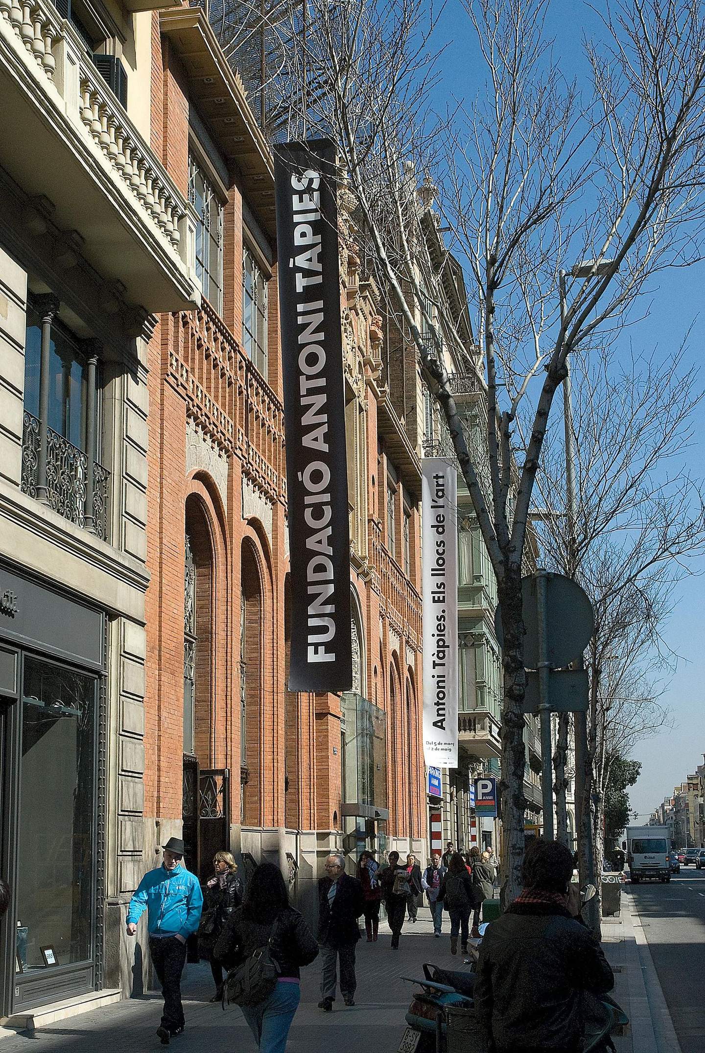 나무와 다양한 상점들이 늘어선 번화한 거리에 있는 안토니 타피에스 재단 (Fundació Antoni Tàpies) 건물 옆을 사람들이 지나갑니다.