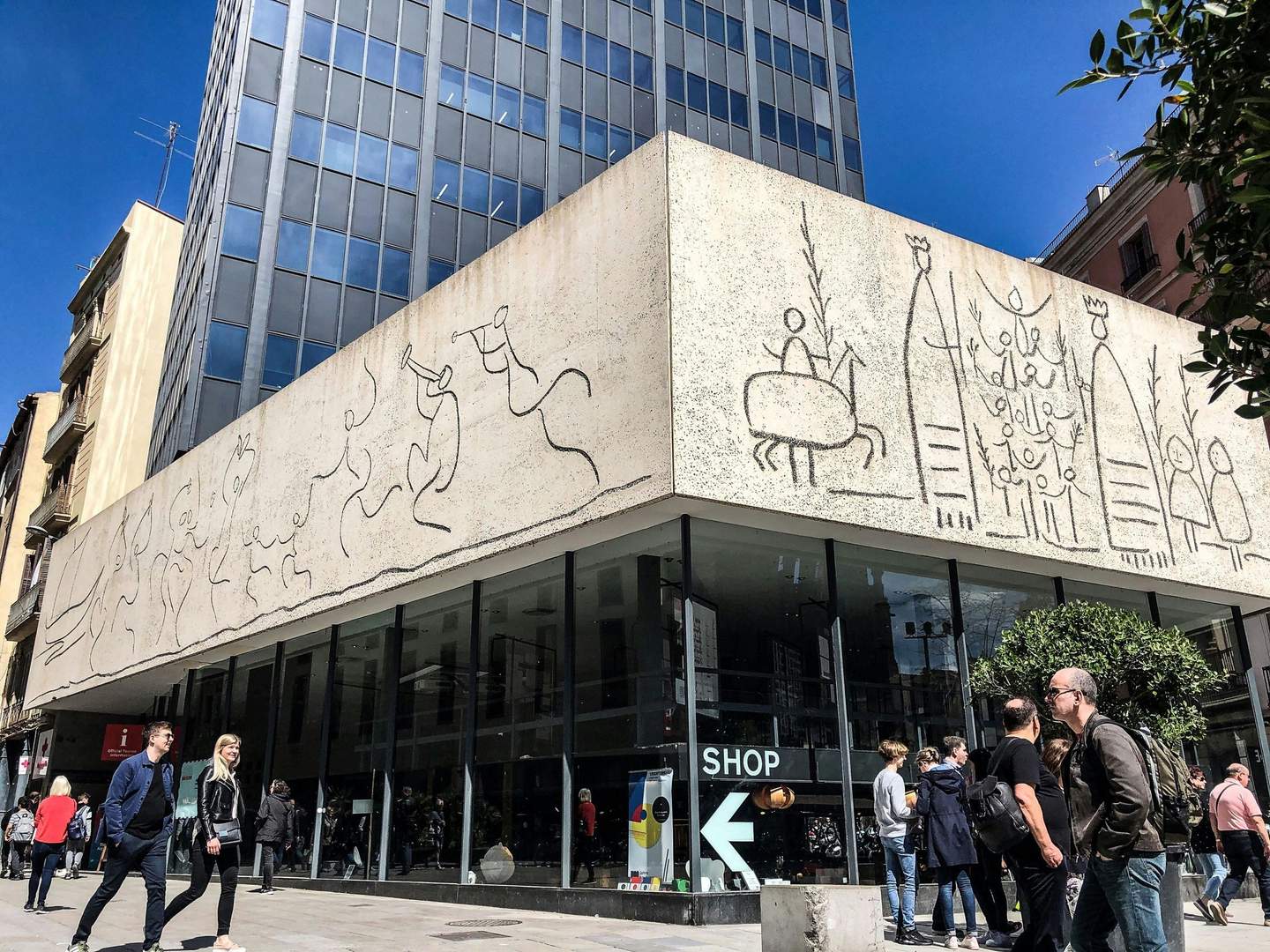 Un edifici modern amb dibuixos de línies a la façana, gent passant i una botiga amb vidre a peu de carrer.