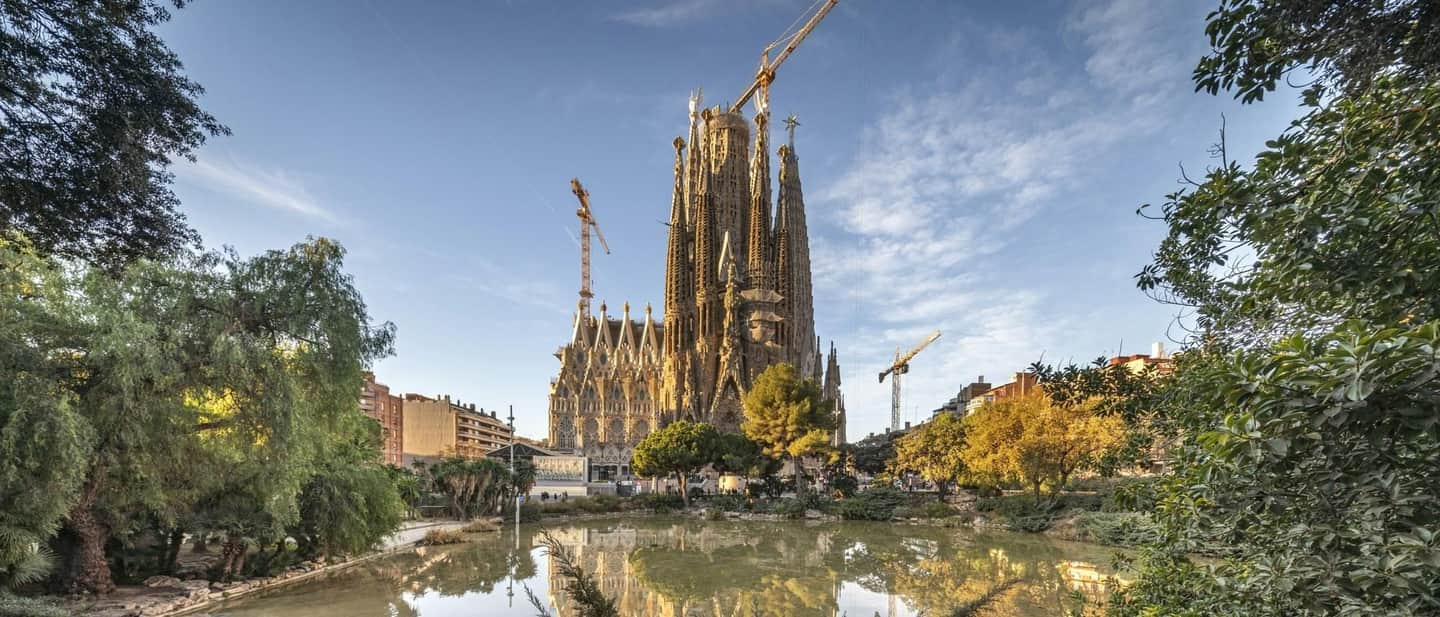 Sagrada Familia The image shows a large, unfinished cathedral with cranes, viewed from a park with lush trees and a reflective pond.