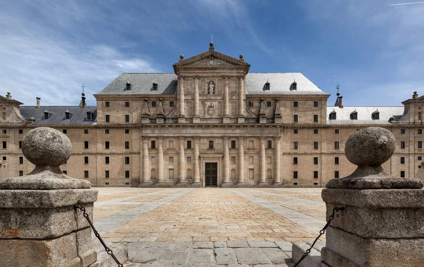 Royal Site of San Lorenzo de El Escorial City Card (Madrid) A historic building with a symmetrical facade, columns, and a central statue, framed by two stone pillars, under a blue sky.