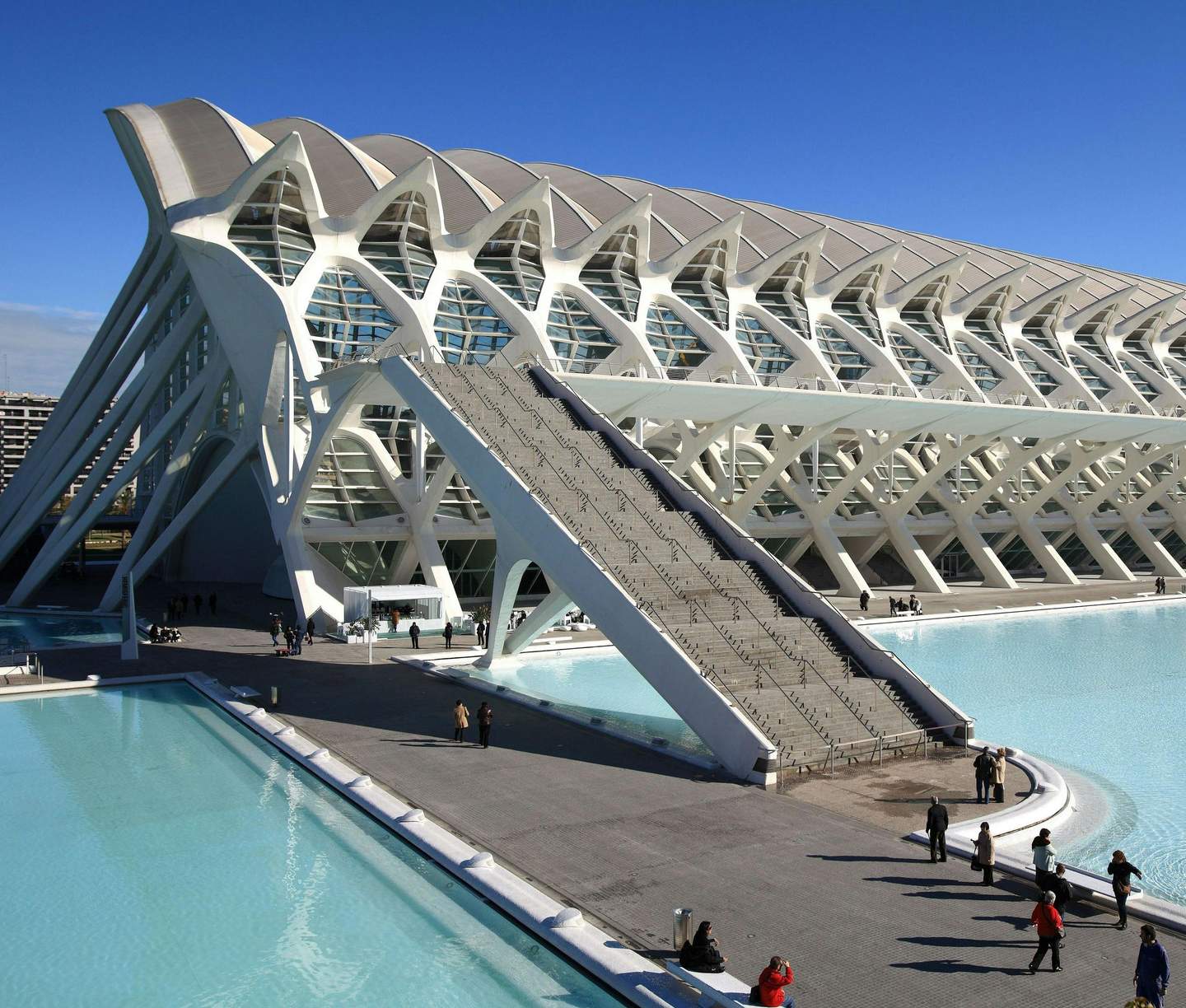 Modern white building with an intricate design, adjacent to reflective pools of water; people walking and gathering nearby.
