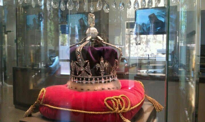 A royal crown with jewels displayed on a red velvet cushion inside a glass case, surrounded by reflective surfaces and lights.