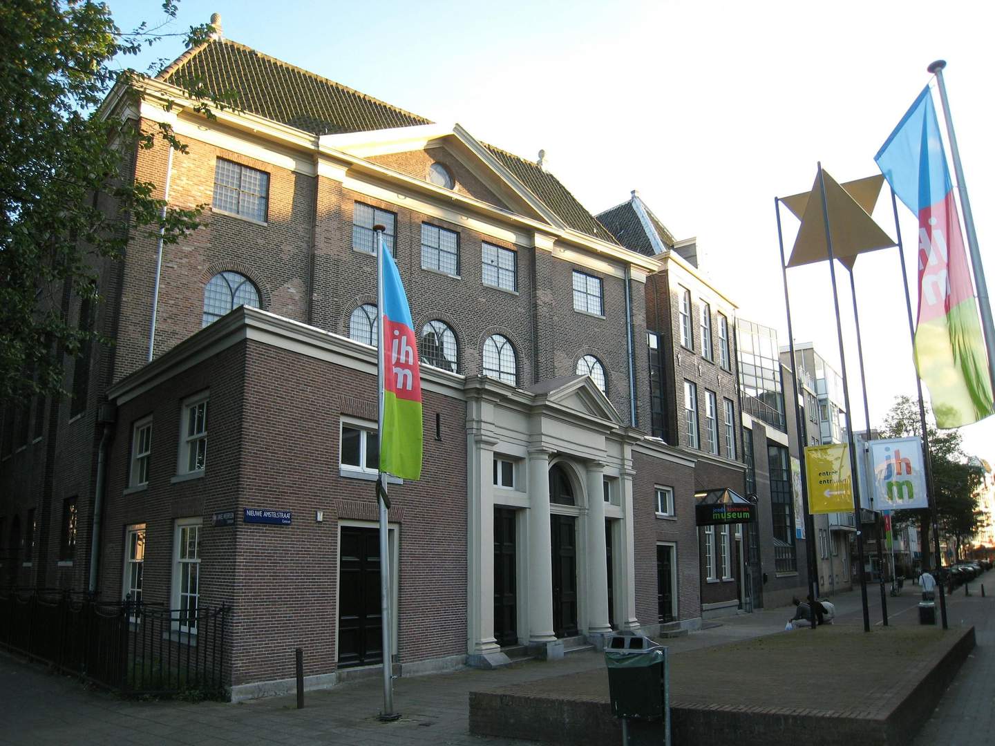 Brick building with arched windows, flags, and "JHM" museum signs. Adjacent sidewalk with trees and benches.
