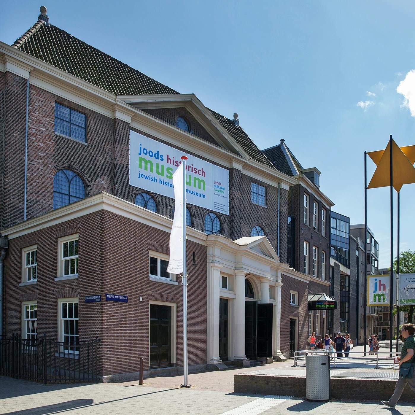 Red-brick building with a sign reading "Jewish Historical Museum" in front, people walking around, and a blue sky above.