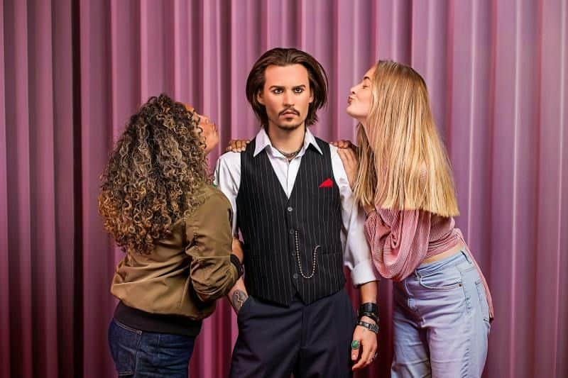 Madame Tussauds Amsterdam City Card (Amsterdam) Three people pose in front of a pink curtain; a man in a vest stands between two women, one on each side, both leaning in toward him.