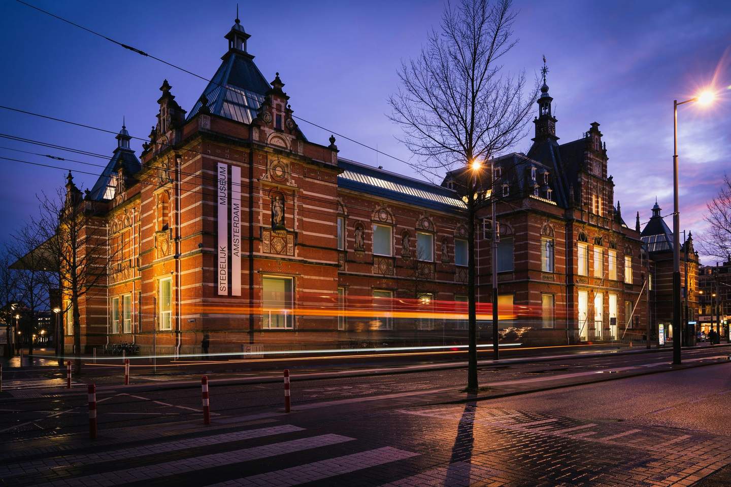 Stedelijk Museum Amsterdam City Card (Amsterdam) Historic building with ornate architecture and a sign reading "Stedelijk Museum Amsterdam" at dusk, with light trails from passing traffic.