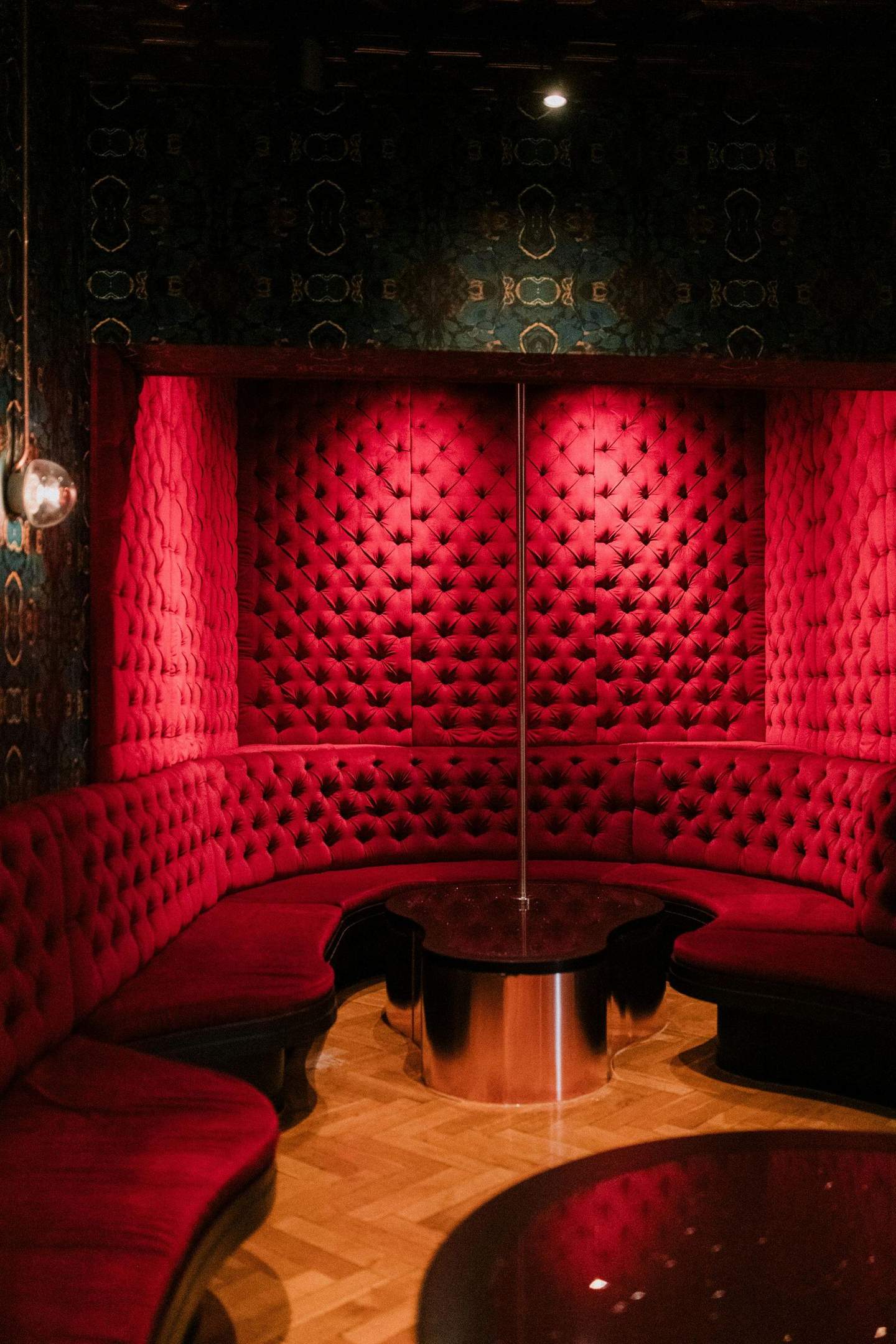 The BonTon Tour City Card (Amsterdam) A dimly lit room with red, tufted velvet seating arranged in a semicircle around a pole. There's a round black table in the center.