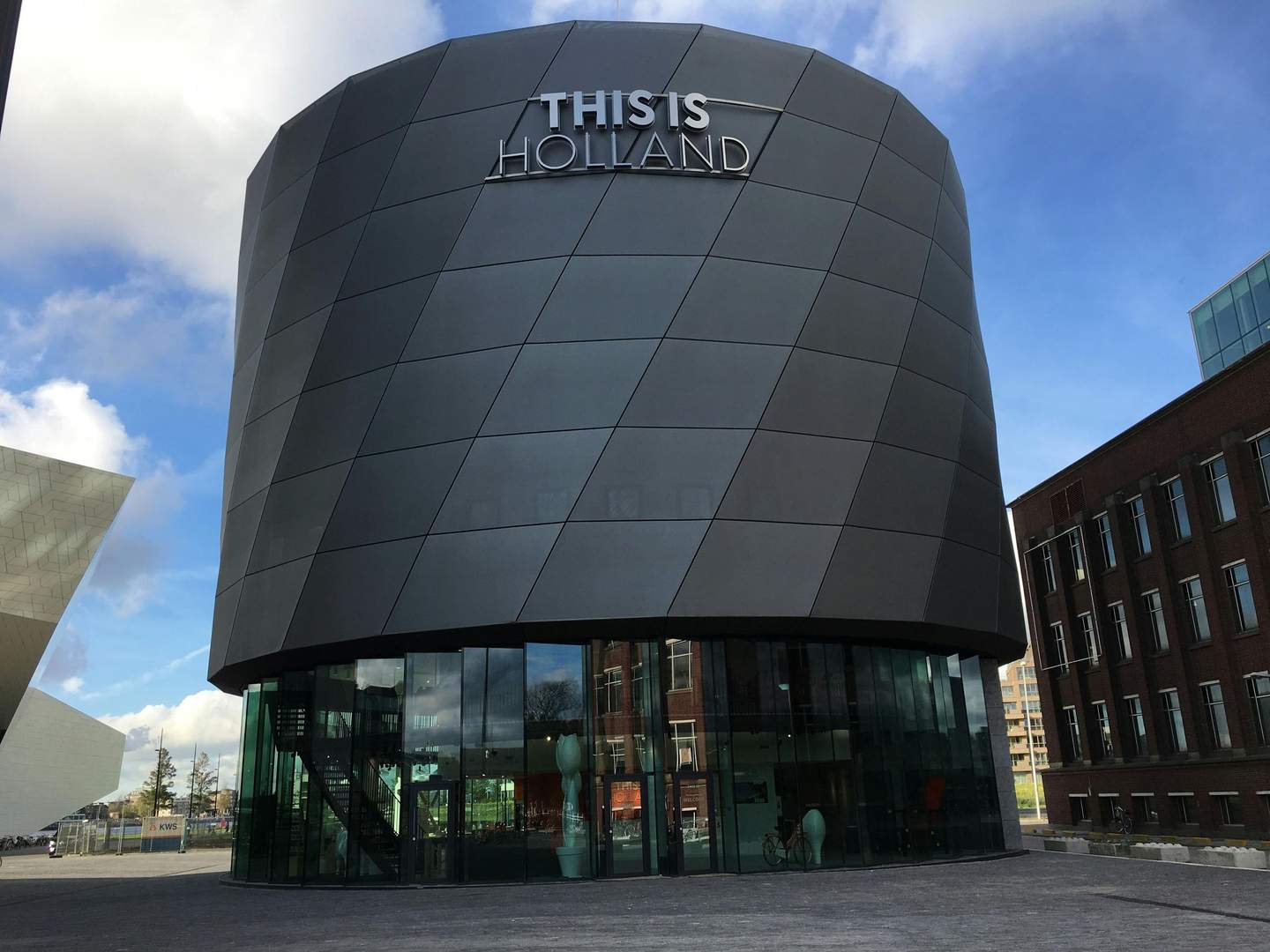 Round, modern building with "THIS IS HOLLAND" sign against a blue sky, with nearby structures and glass entrance at ground level.