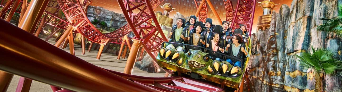 IMG Worlds of Adventure City Card (Dubai) People riding a rollercoaster with a dinosaur-themed front car, surrounded by a red track structure and artificial rock formations.