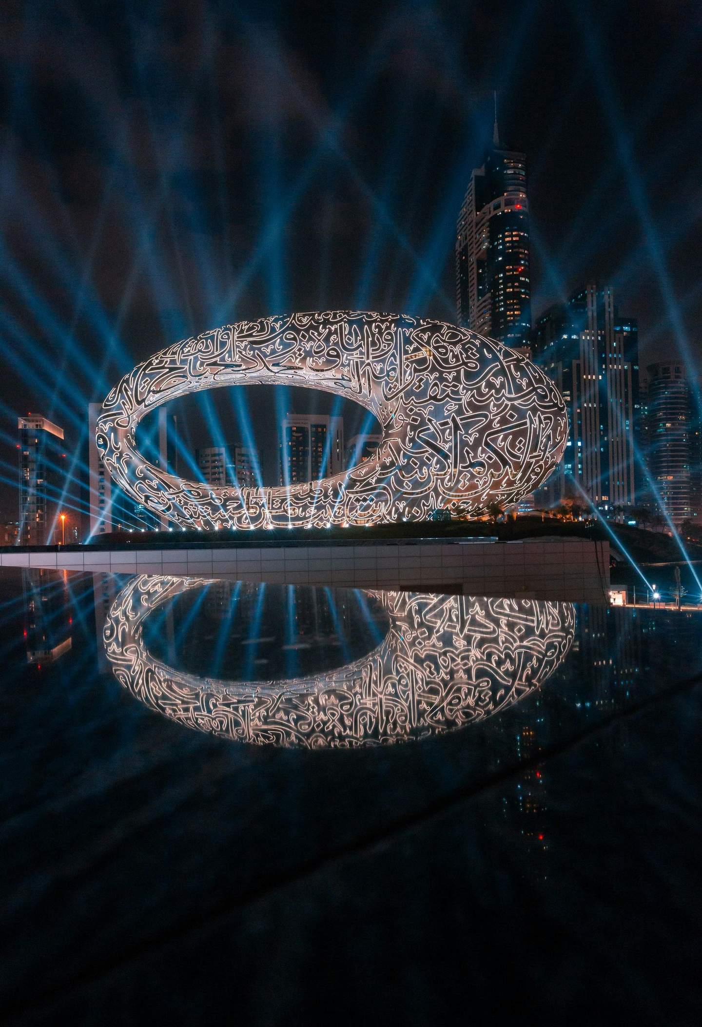 A futuristic oval-shaped building with glowing script patterns, set against a cityscape with skyscrapers and illuminated by blue beams.