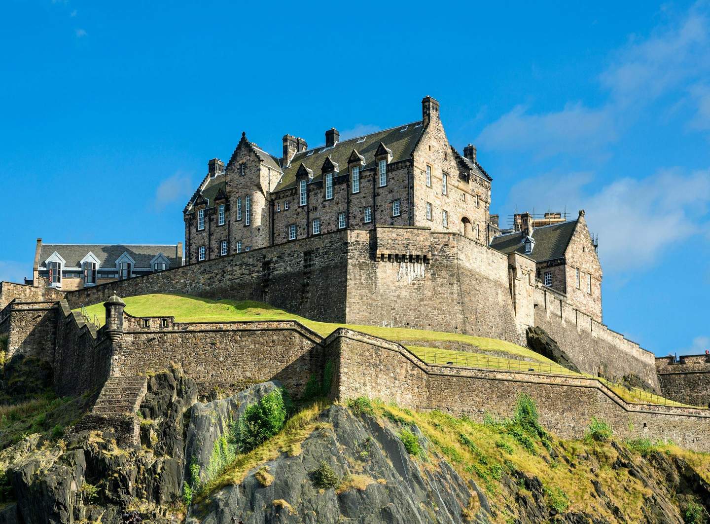 Historic stone castle on a rocky hilltop under a clear blue sky, featuring multiple connected buildings and sloped grassy areas.