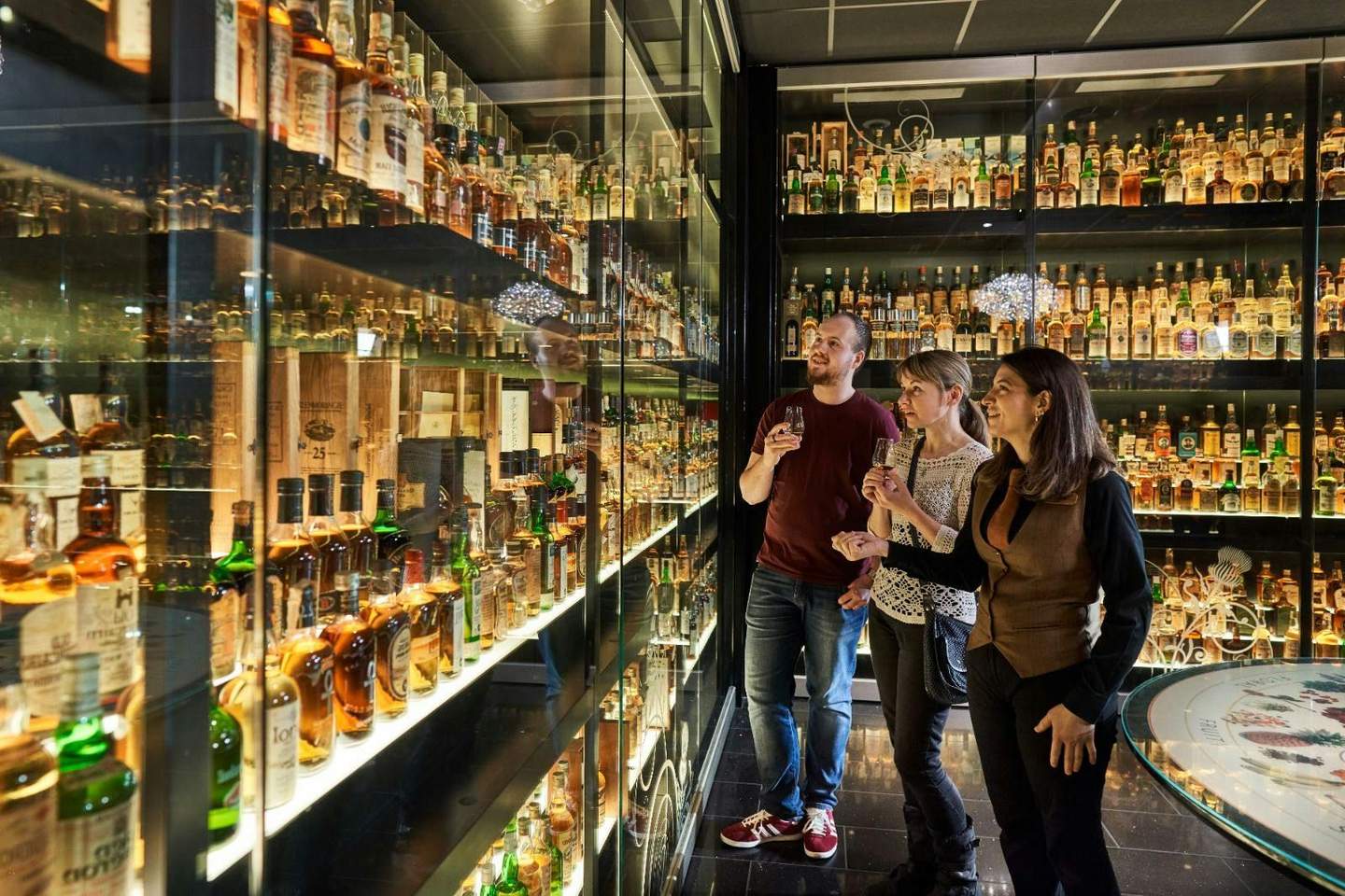 The Scotch Whisky Experience City Card (Edinburgh) Three people examining shelves filled with numerous bottles of whiskey in a well-lit display room.