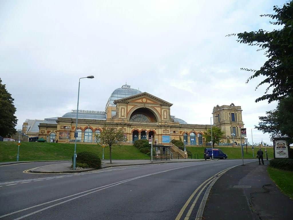 A large historic building with an arched entrance, flanked by towers, seen from the street with a van and a few pedestrians.