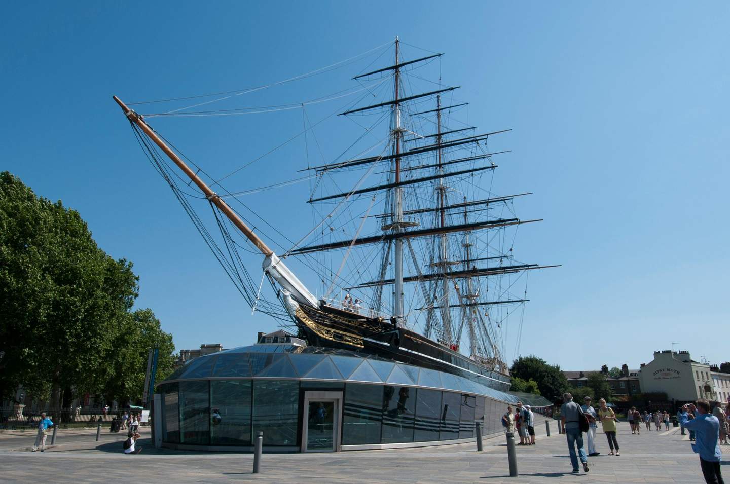 A historic ship with tall masts and a bowsprit docked on a glass-encased platform, with people walking and trees nearby.