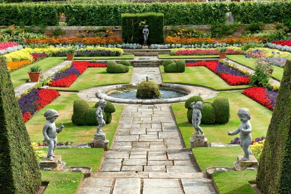 Hampton Court Palace A formal garden with colorful flower beds, stone pathways, a central fountain, and statues of children positioned symmetrically.