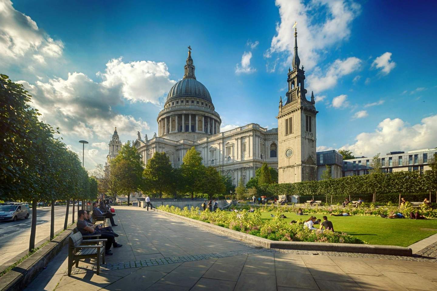 People relaxing on benches and grass near a historic white-domed building with towers, under a sky with scattered clouds.