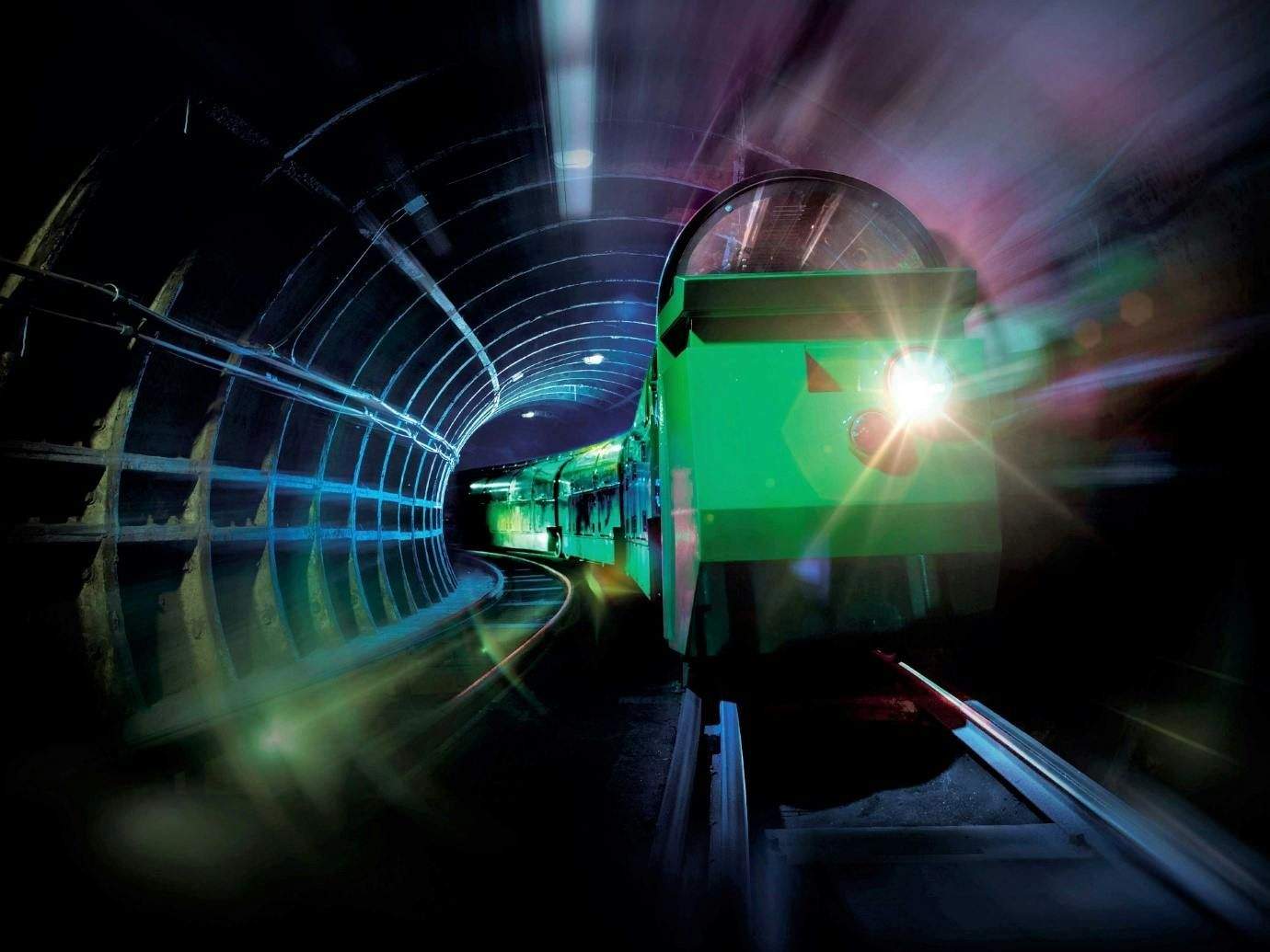 Un tren verd amb una llum frontal brillant viatja a través d'un túnel poc il·luminat amb parets corbes. Els senders de llum emfatitzen el moviment.