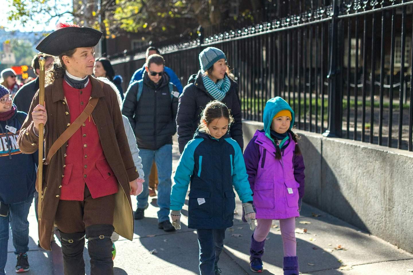 A person in historical attire leads a group of people, including children in colorful winter coats, walking on a sidewalk.