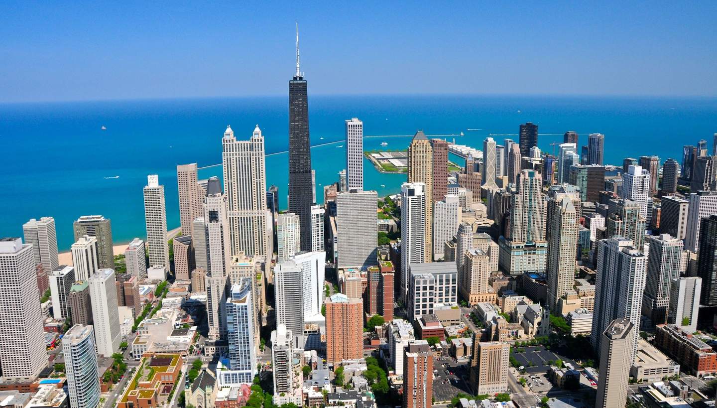 Aerial view of a city with numerous tall buildings near a large body of water under a clear blue sky.