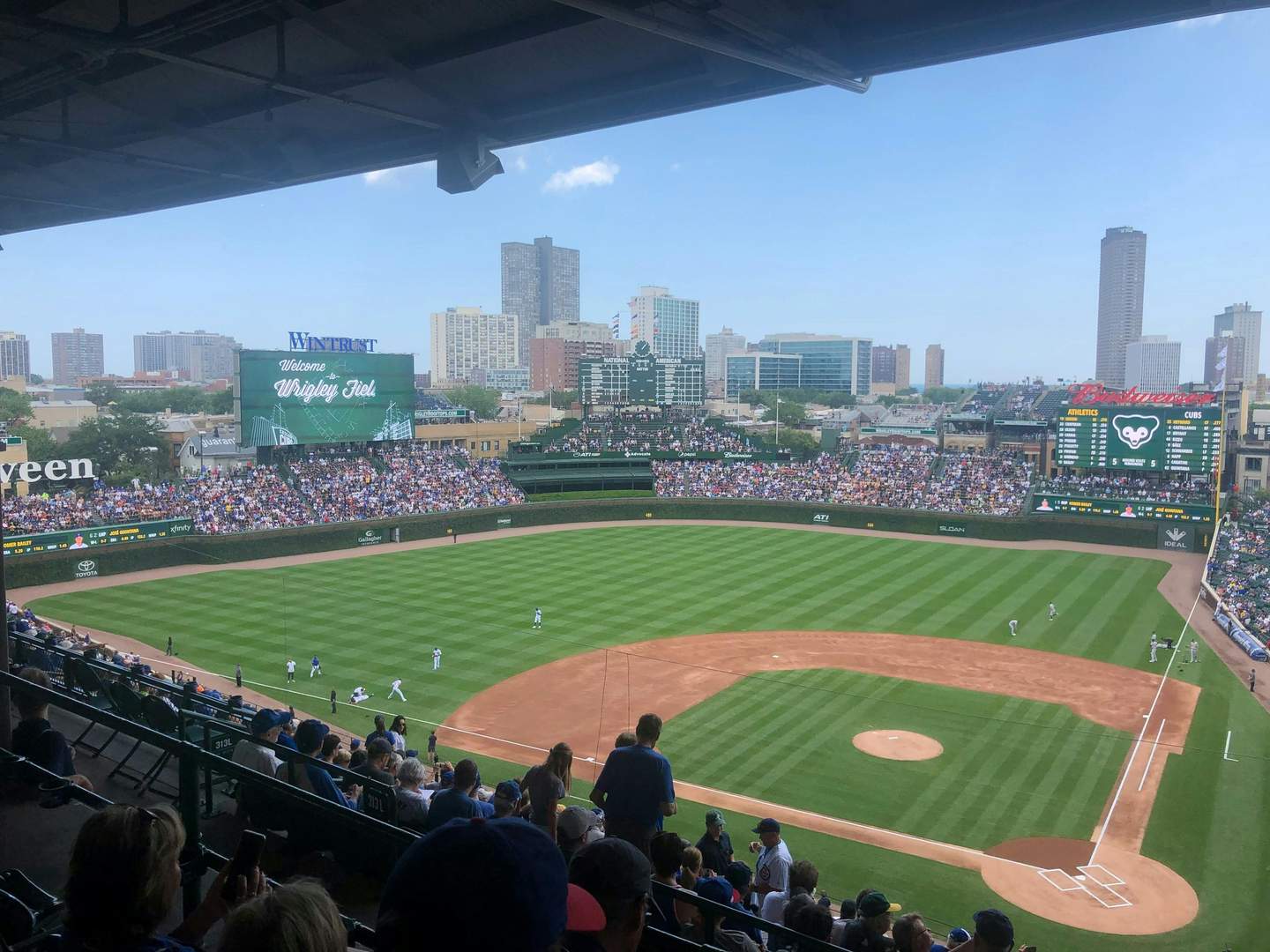 Wrigley Field during a baseball game, crowded stands, players on the field, and the scoreboard displaying "Welcome to Wrigley Field."