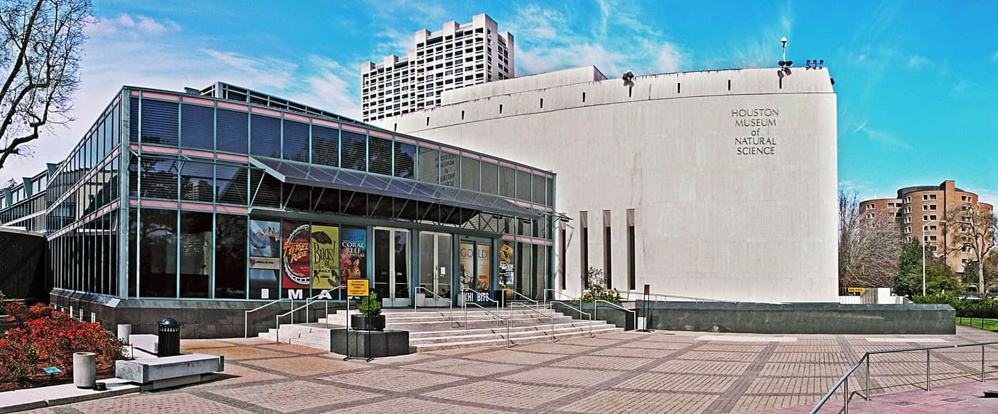 Natuurhistorisch Museum Houston Modern gebouw met glazen gevel en toegangsposters voor tentoonstellingen in het Houston Museum of Natural Science. Nabijgelegen hoogbouw op de achtergrond.