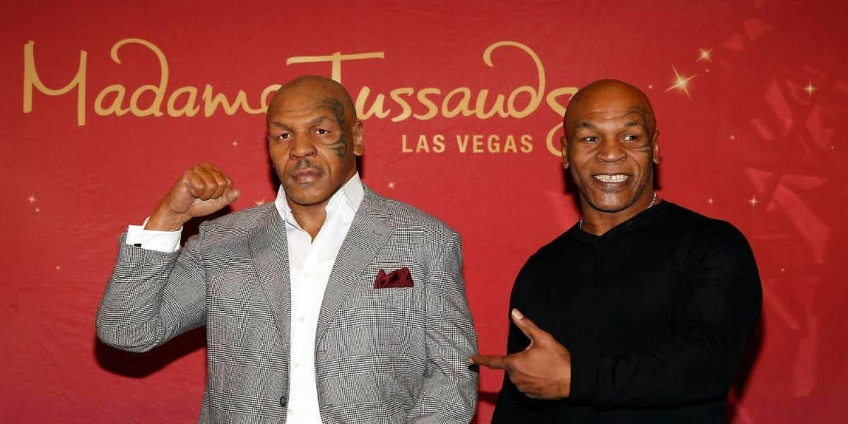 Two men stand in front of a Madame Tussauds Las Vegas backdrop. One man is wearing a suit, and the other is in a black shirt.