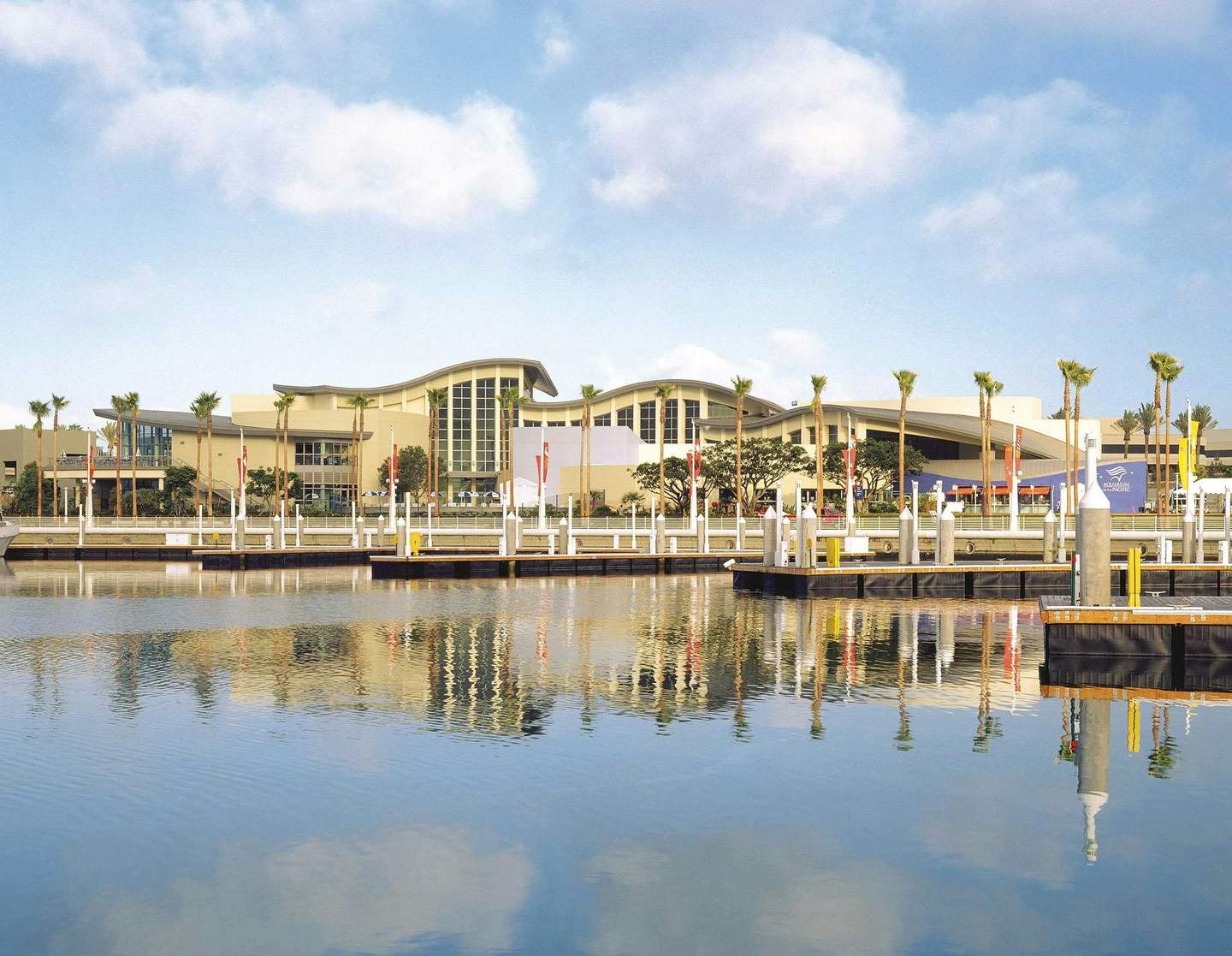 Aquarium of the Pacific City Card (Los Angeles) A waterside view of a modern building with palm trees, docks, and calm water reflecting the scene under a partly cloudy sky.