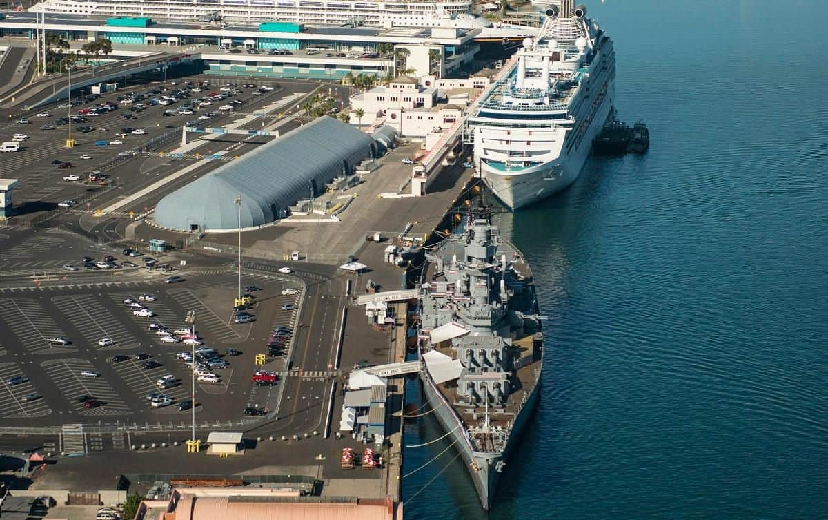 Aerial view of a cruise ship and a battleship docked at a port with adjacent parking lots and terminal buildings.